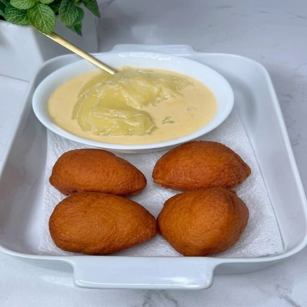 Four golden fried dough balls and a bowl of creamy yellow pap with a spoon on a white tray.